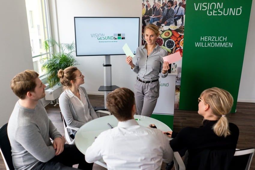 Es ist ein Meetingraum zu sehen. Vier Personen sitzen an einem runten Tisch und hören einer Präsentation zu, welche von einer Frau gehalten wird.