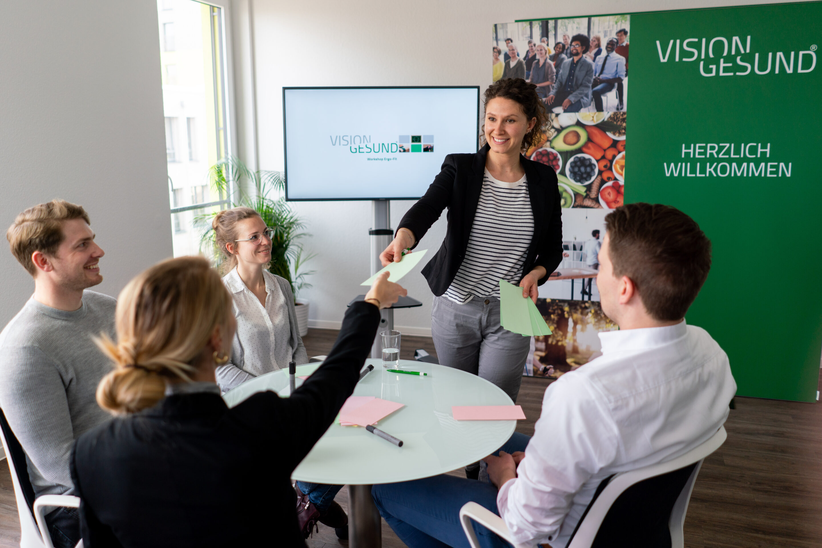 Mehrere Personen sitzen um einen runden Tisch in einem modernen Besprechungsraum, während eine weitere Person vorne steht und eine Präsentation oder Besprechung leitet.