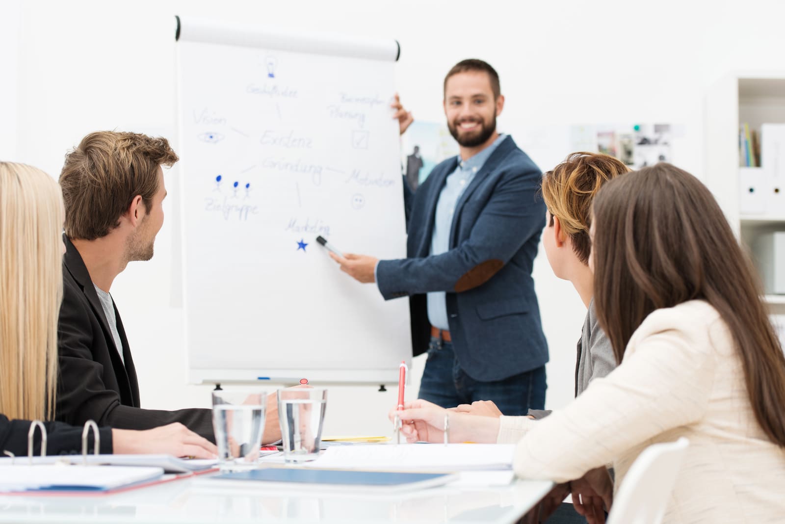 Workshop Gesunde Führung Eine Person präsentiert Inhalte auf einem Whiteboard vor einer kleinen Gruppe in einem modernen Besprechungsraum. Mehrere Teilnehmende sitzen am Tisch, schauen auf die Präsentation und haben Unterlagen sowie Getränke vor sich.