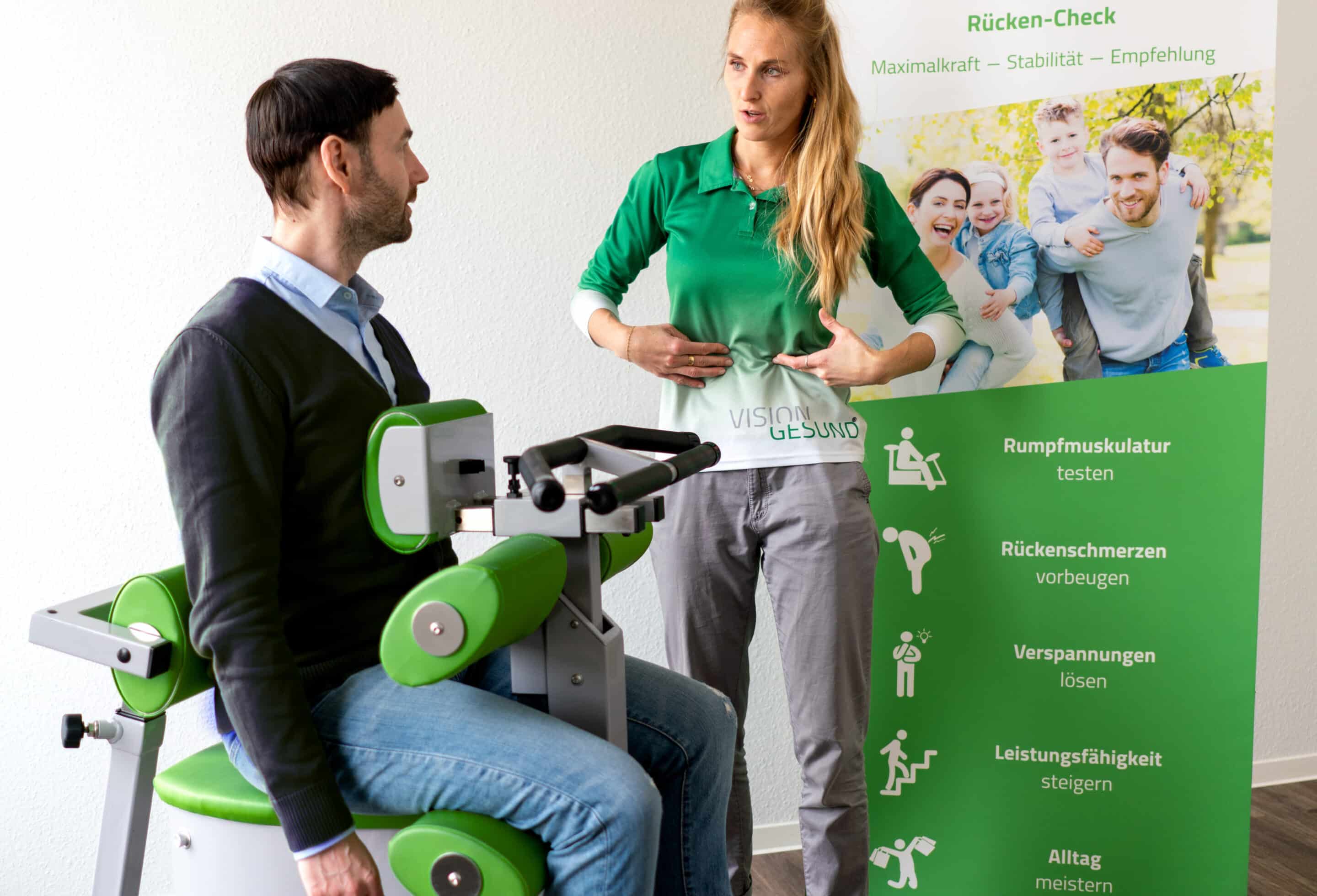 Auf dem Bild sieht man eine Frau, die bei einem Mann den Rücken-Check durchführt. Dafür sitzt der Mann an einem Sportgerät. Die Frau steht neben dem Mann und gibt ihm Anweisungen.
