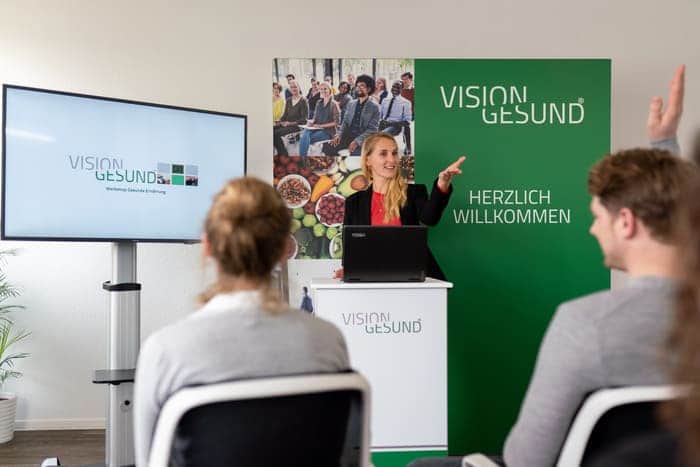 Eine Vortragssituation bei VisionGesund, in der eine Person am Rednerpult präsentiert und Teilnehmende im Raum zuhören und Fragen stellen. Neben dem Podium stehen ein Bildschirm mit dem VisionGesund‑Logo und eine grüne Willkommenswand.