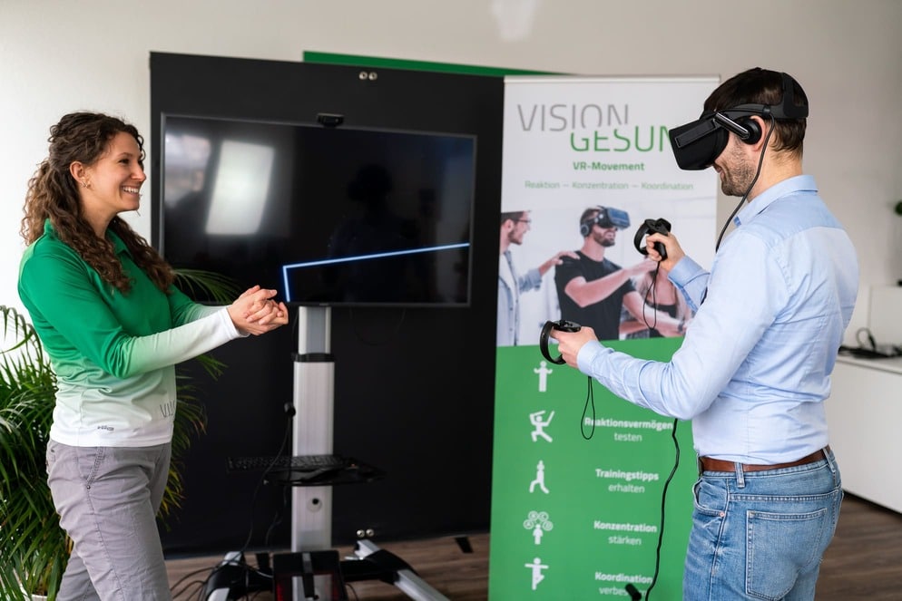 Ein Mann hat eine VR Brille aufgesetzt und führt Bewegungen aus. Dabei steht eine Frau daneben und leitet ihn an.