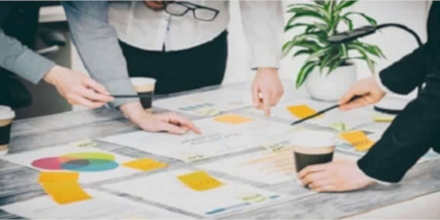 Ein Tisch mit vielen Zetteln ist zu sehen. Auf diesen Zetteln sieht man Statistiken oder Grafiken. Hände von 3 Personen sind zu sehen, die mit Stiften in ihren Händen oder mit ihren Fingern auf die Zettel zeigen.