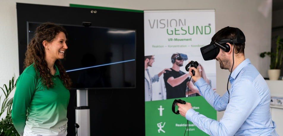 Eine Person bedienen ein Virtual-Reality-System für Bewegungs- oder Ergonomietraining. Auf einem Bildschirm im Hintergrund wird eine VR-Anwendung angezeigt.