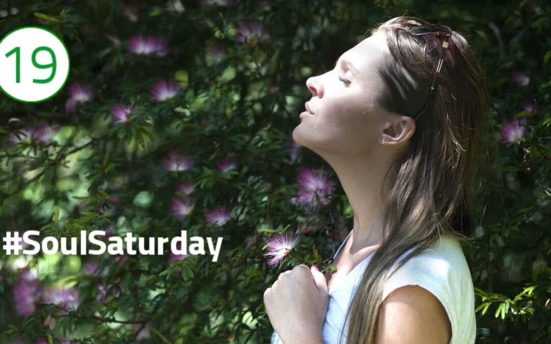 Eine Frau reckt mit geschlossenen Augen ihr Gesicht in die Sonne, die von der linken Bildseite einfällt. Im Hintergrund sind lila Blüten in einem Busch zu sehen, was dem Bild einen Ausdruck der Naturverbundenheit, Ruhe und Stille verleiht.