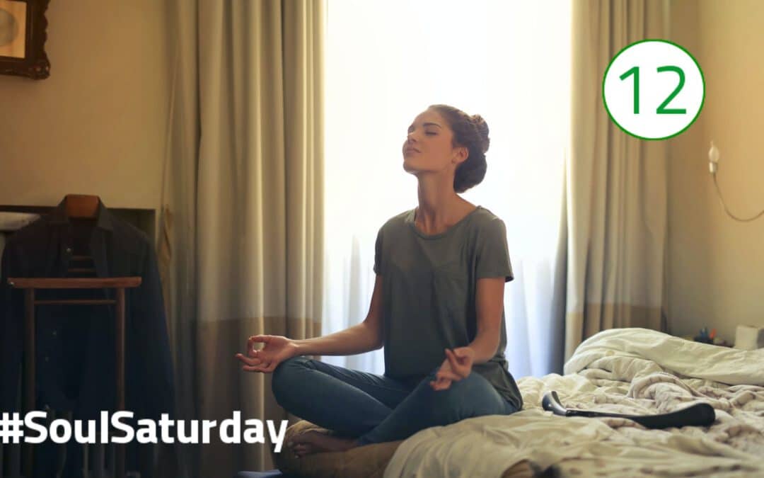 Eine Frau sitz im T-Shirt und Jeans im Schneidersitz auf der Bettkante und meditiert. Die Augen sind dabei geschlossen und der Kopf zur Decke gerichtet.